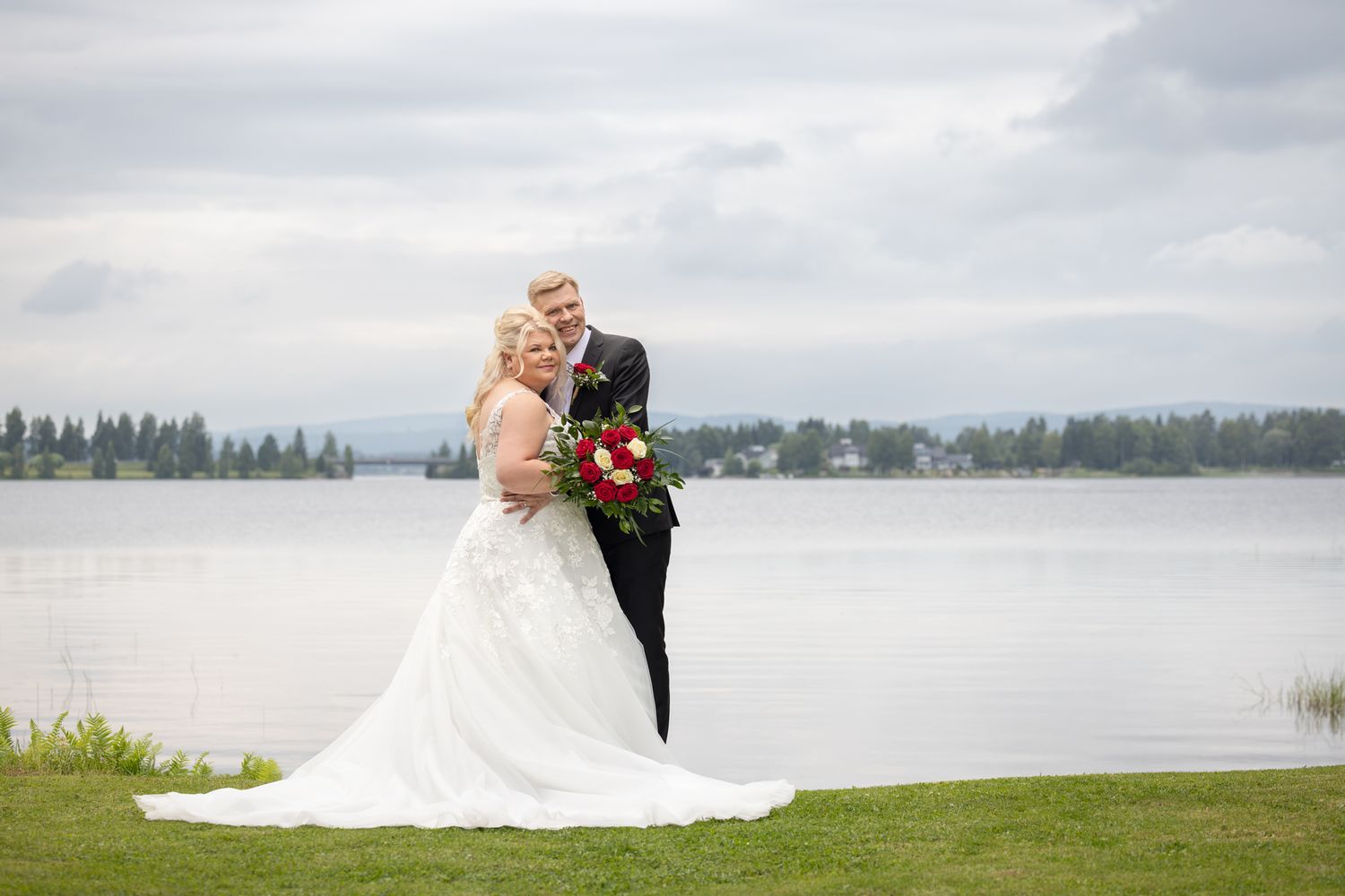 Bride and groom posing by a serene lakeside on their wedding day, bride holding a red bouquet in a white gown.