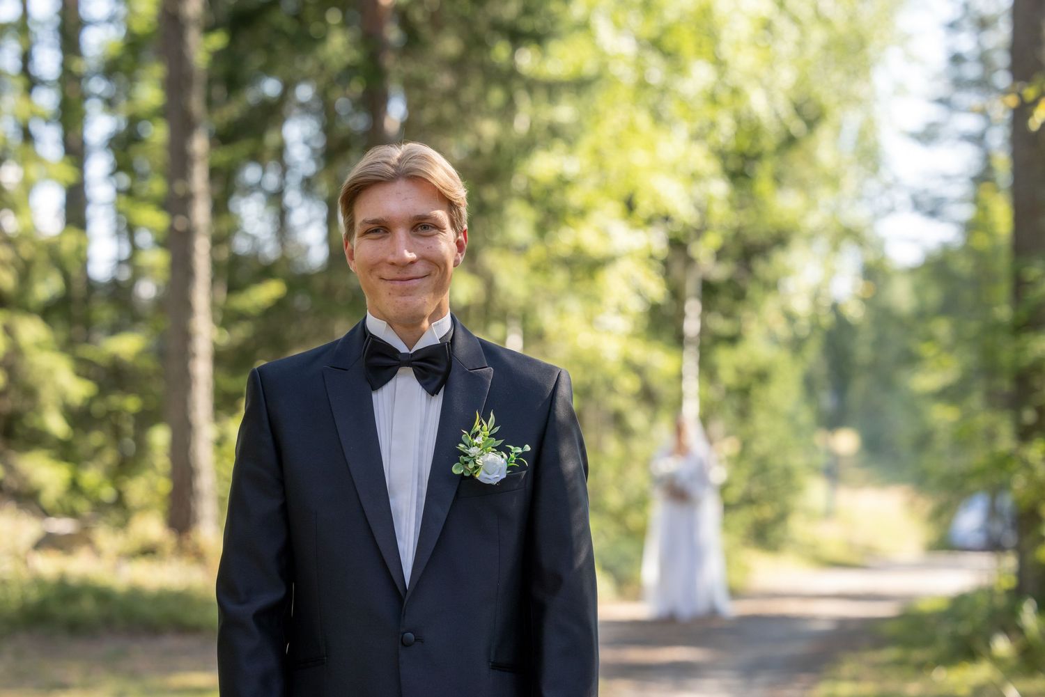 Smiling groom in black tuxedo with boutonniere stands on forest path on his wedding day.
