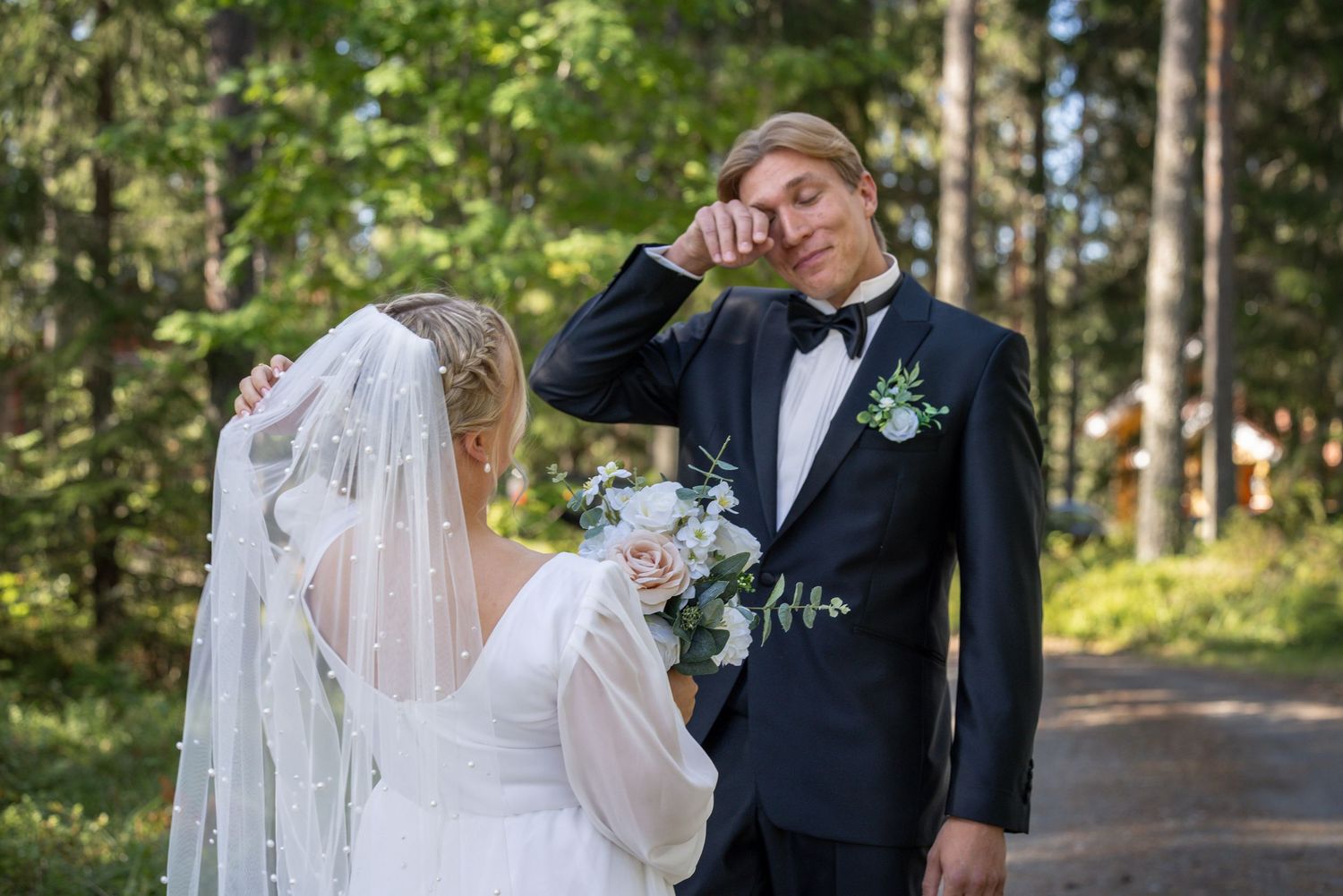 Emotional groom wiping tears in a black tuxedo beside his bride in white dress and veil, holding bouquet outdoors.