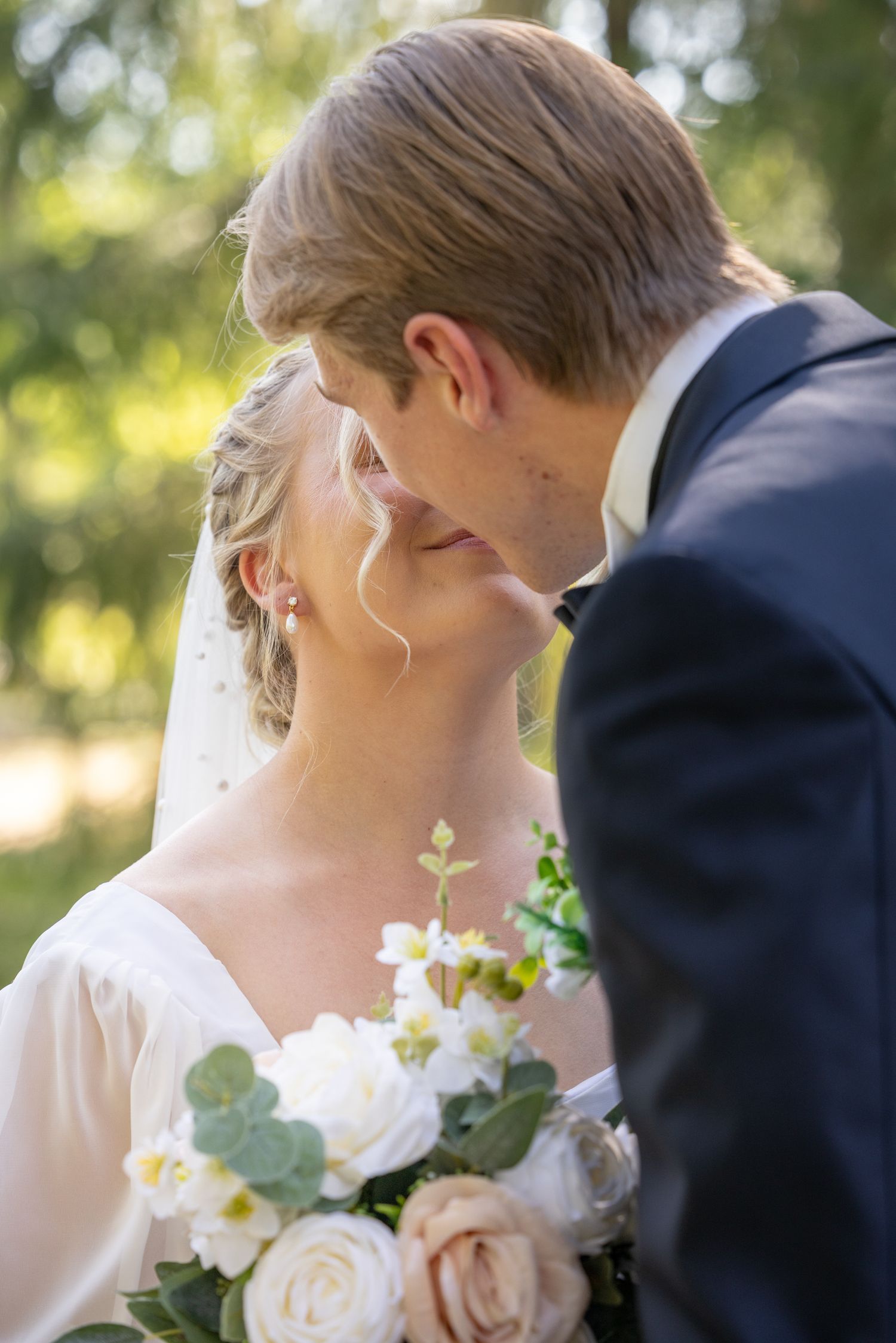 Bride and groom sharing a romantic moment, nearly kissing, with a white floral bouquet in a sunlit outdoor setting.