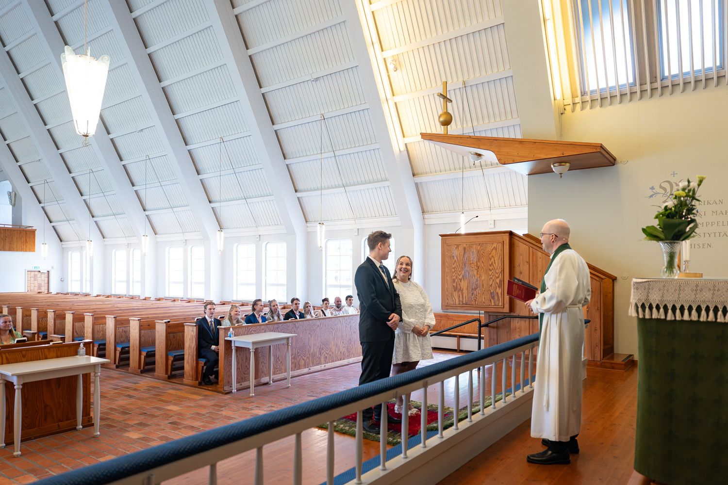 A priest and guest exchange greetings at the altar of a modern church with white vaulted ceilings and wooden pews.
