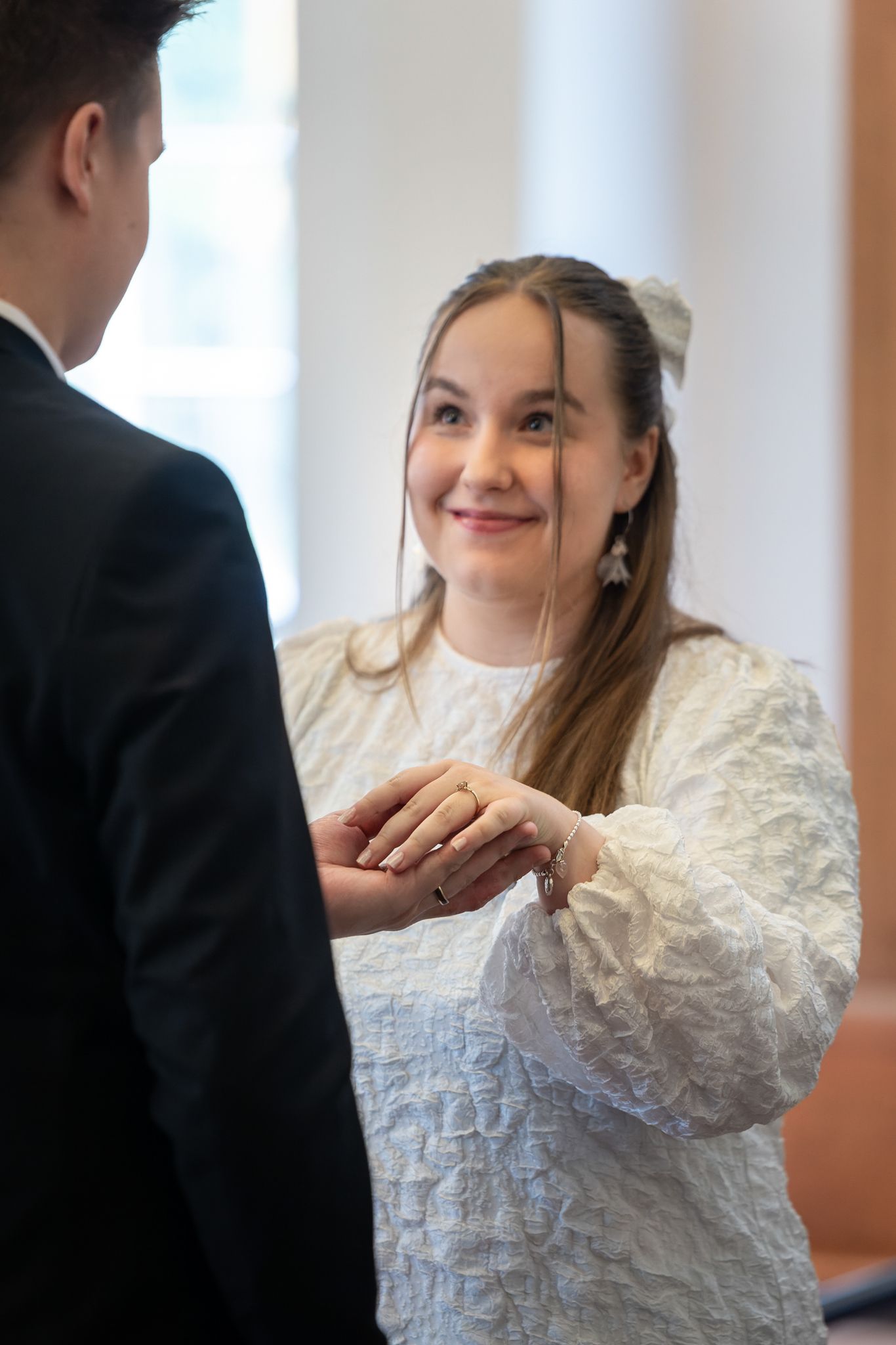 Bride in white dress smiles while exchanging rings with groom in black suit during wedding ceremony.