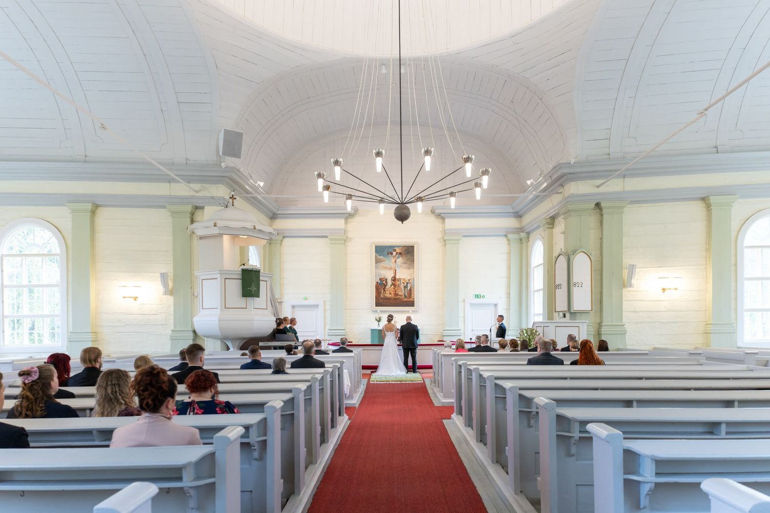 Interior of a traditional white church with wooden pews, red carpet aisle, and elegant chandelier lighting.