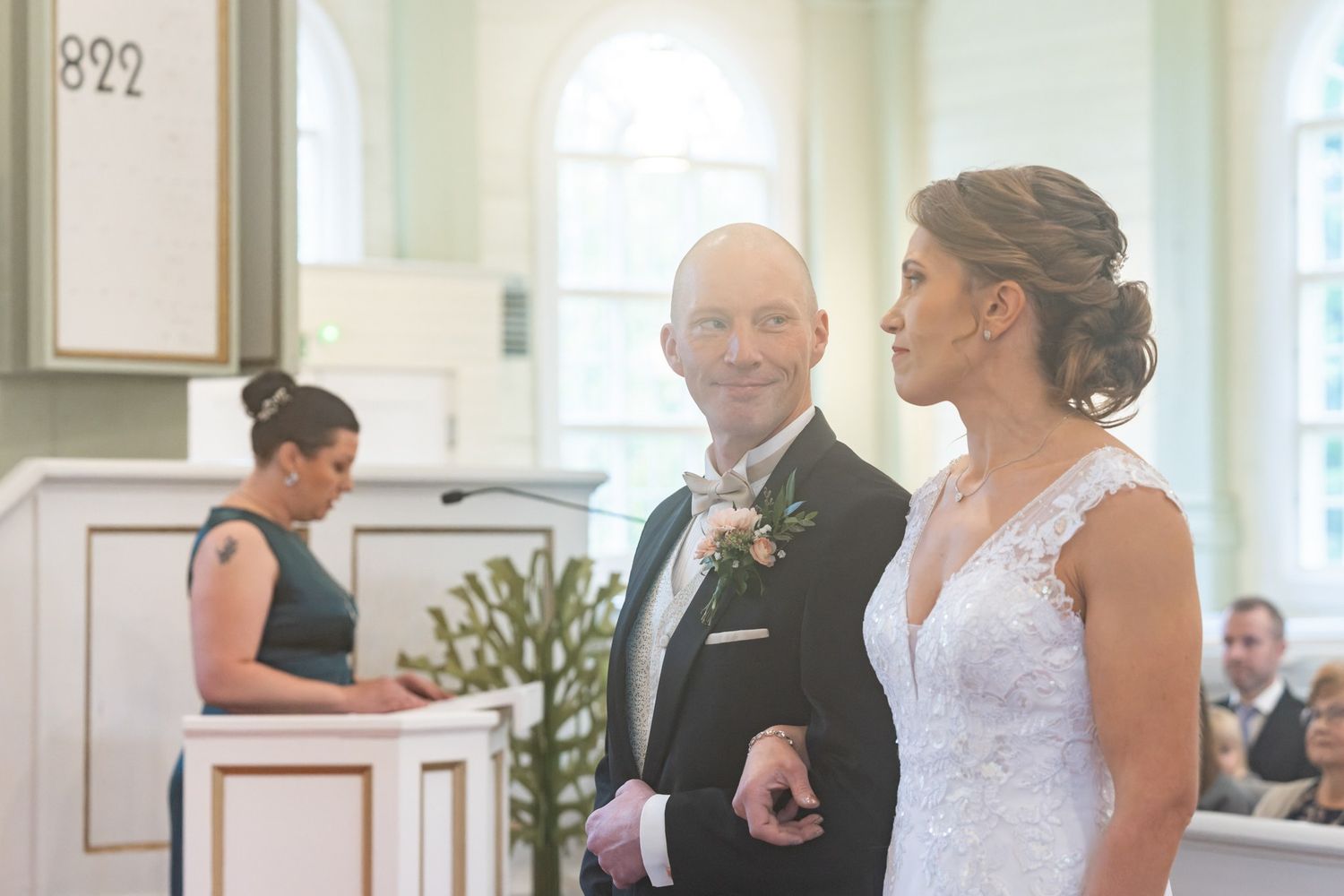 A wedding ceremony takes place in an elegant white room with guests watching from their seats.