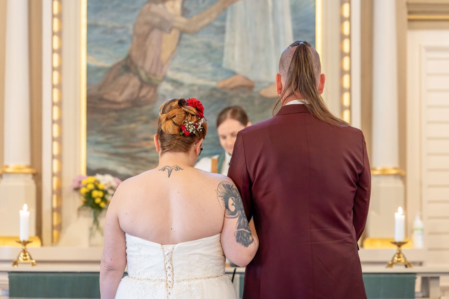 A couple in formal attire stands together during a wedding ceremony in an elegant church with ornate decor.