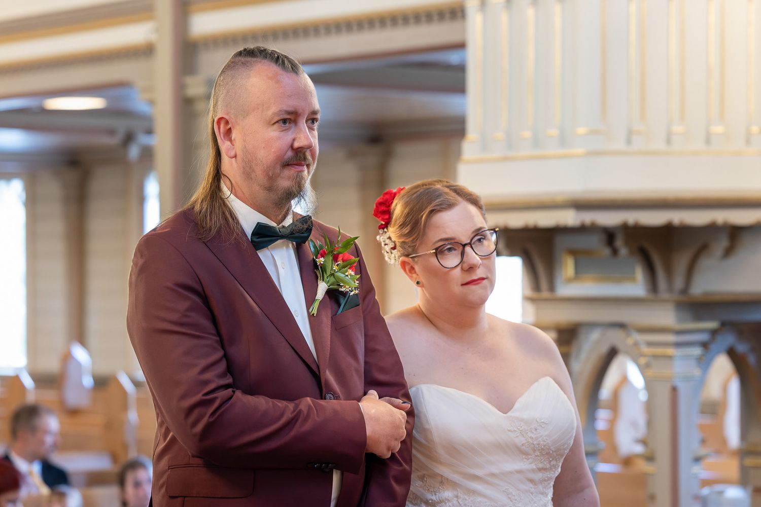 A couple stands together at their wedding ceremony, with the bride in a white dress and groom in a burgundy suit.