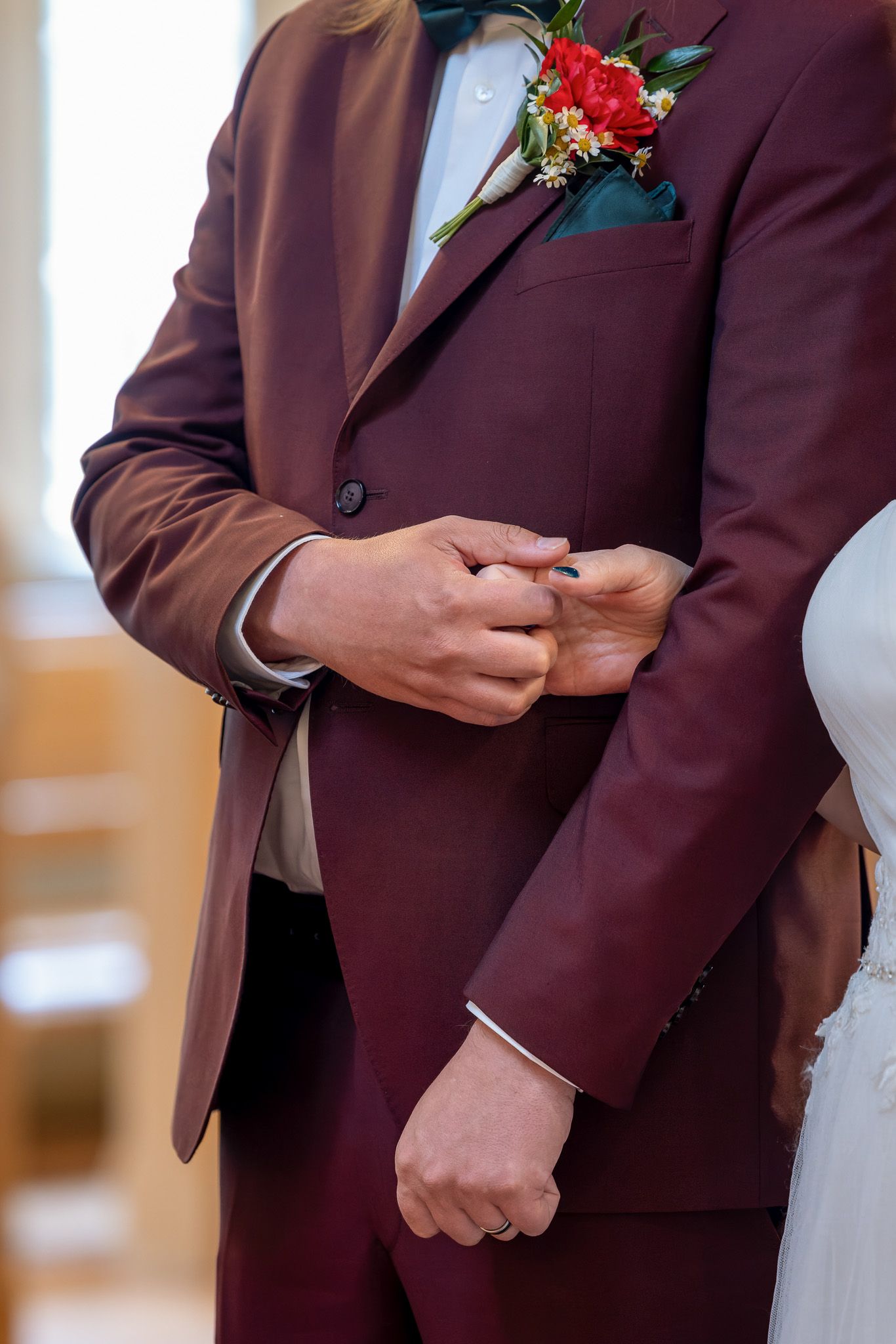 Close-up of a burgundy suit jacket with a red floral boutonniere and hands adjusting the buttons at a formal event.
