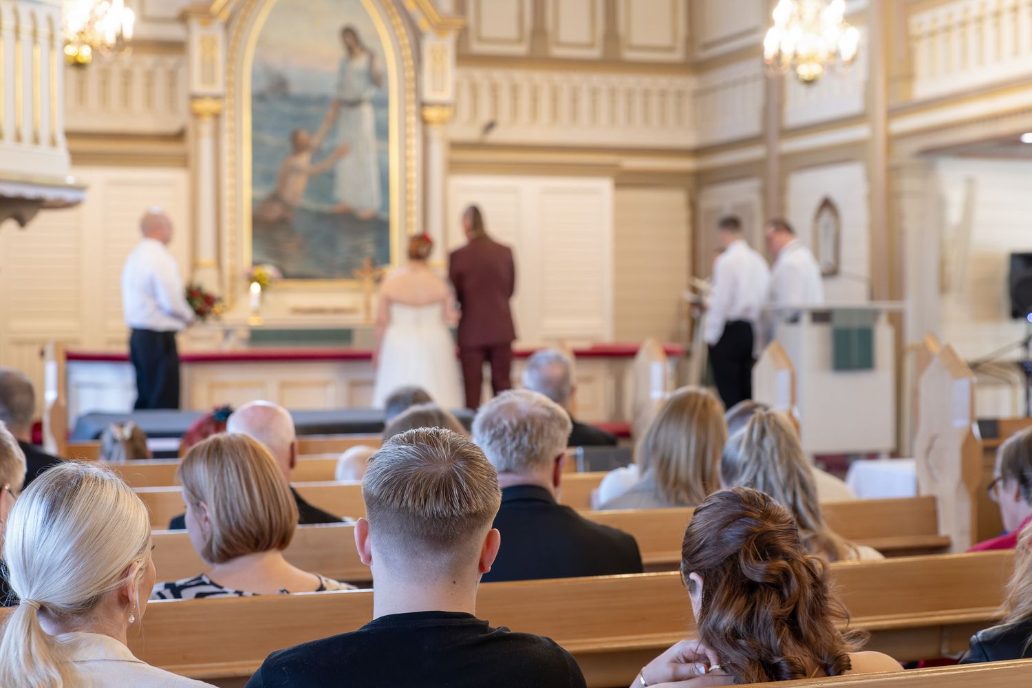 People seated in wooden pews inside an ornate church with elegant chandeliers and decorative architectural details.