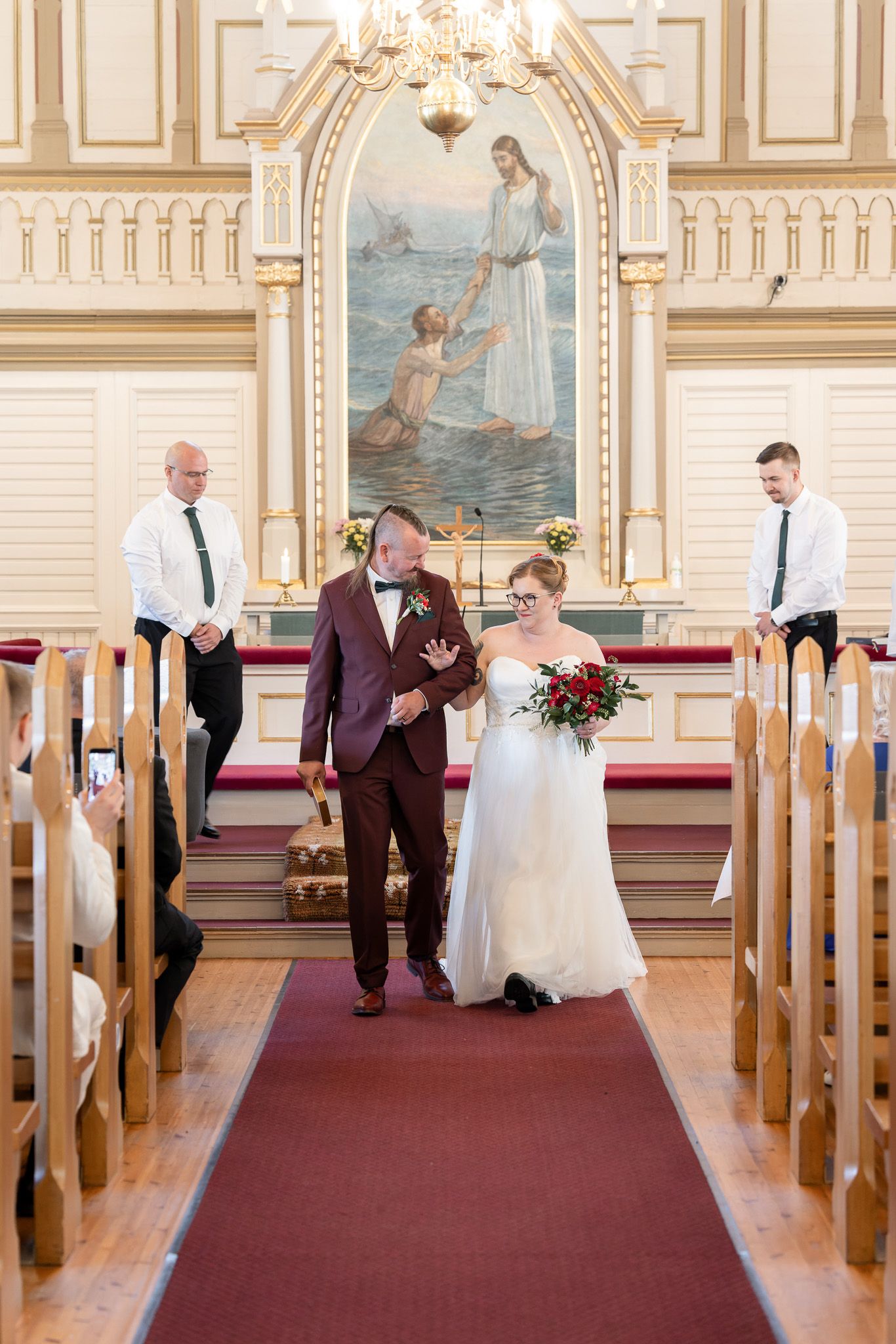 A couple walks down the aisle of a beautiful church with ornate decor and a religious mural on the wall.