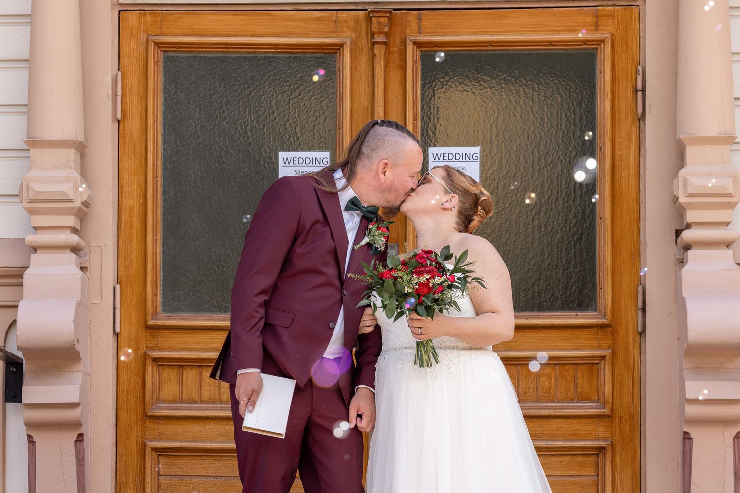 A couple shares a romantic kiss outside wooden doors of an elegant building with ornate pillars.