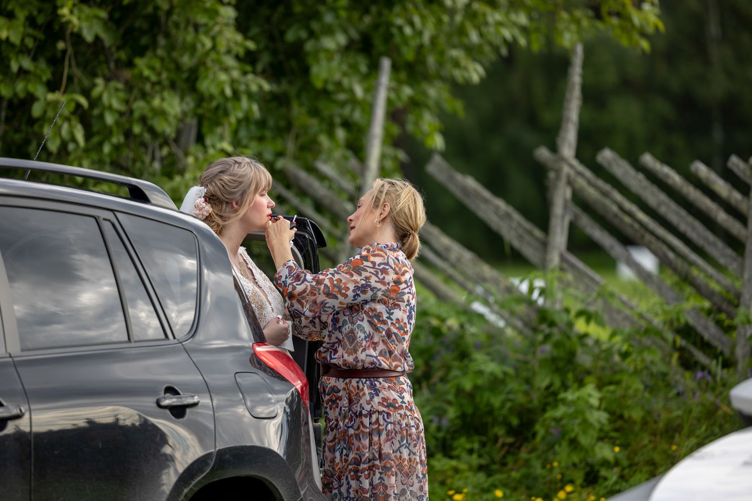 Two women standing beside a dark car outdoors, one applying makeup to the other near a wooden fence.