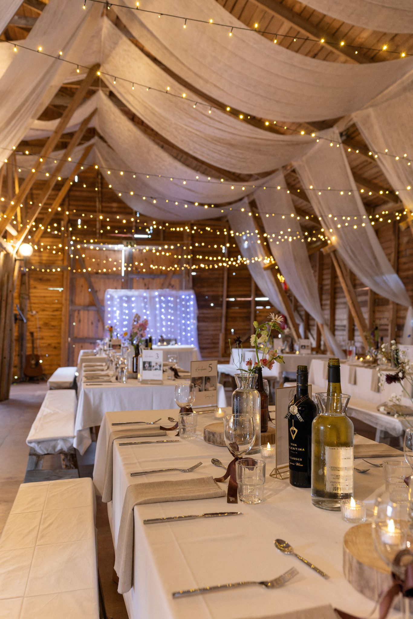 Rustic barn wedding reception with string lights, white draping, set tables, wine bottles, and candles creating an elegant atmosphere.