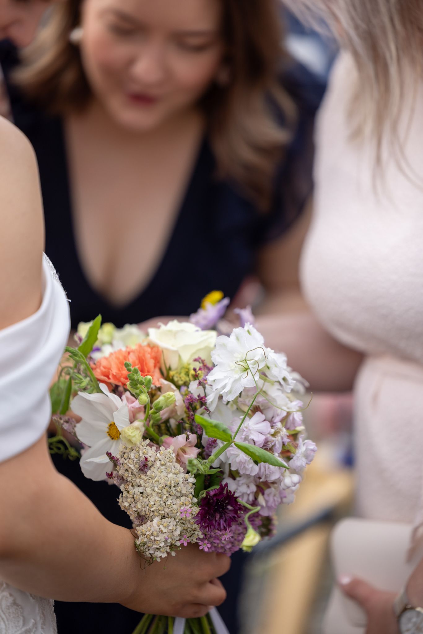Close-up of bride holding colorful wedding bouquet with white, purple, and orange flowers at ceremony.