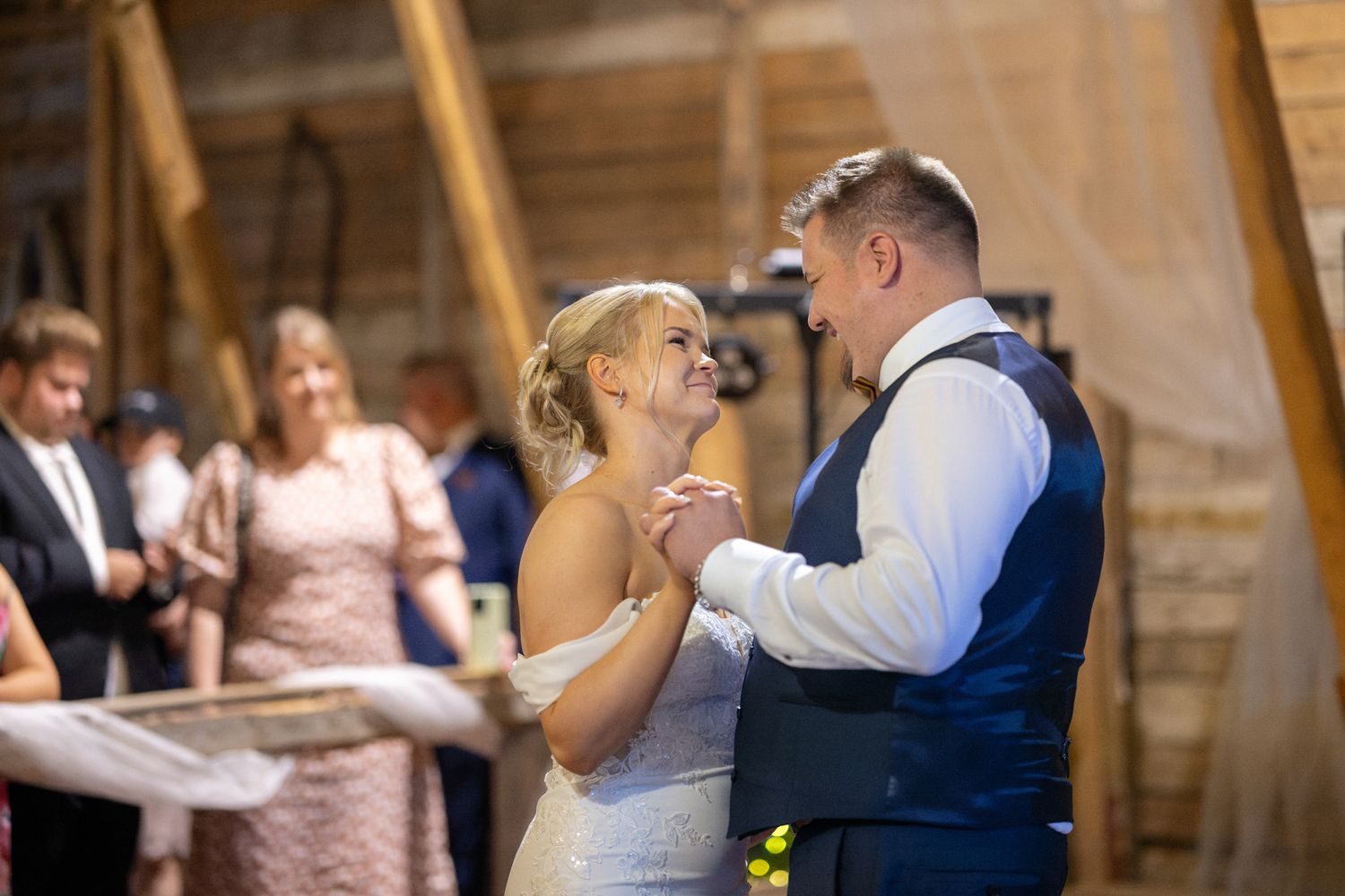 Bride and groom share their first dance in a rustic barn wedding venue, surrounded by smiling guests.