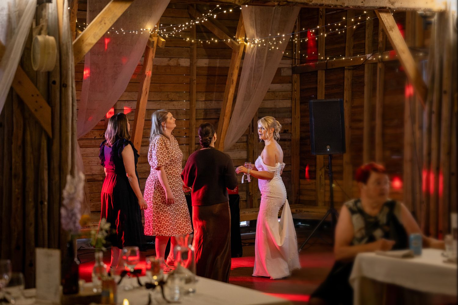 Guests dancing and socializing inside a rustic barn wedding reception with warm string lights and red ambient lighting.