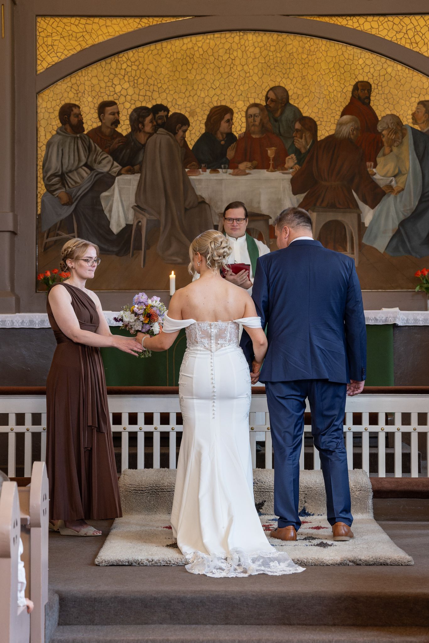 Bride and groom exchanging vows at church altar with Last Supper mosaic backdrop and bridesmaid holding bouquet.