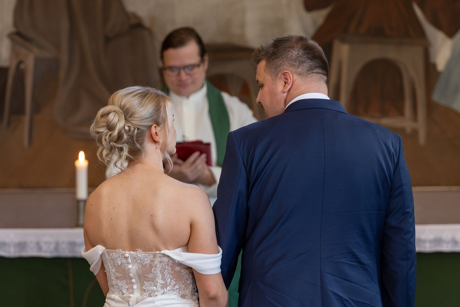 Bride and groom stand before a priest during a church wedding ceremony, with candles and altar in the background.