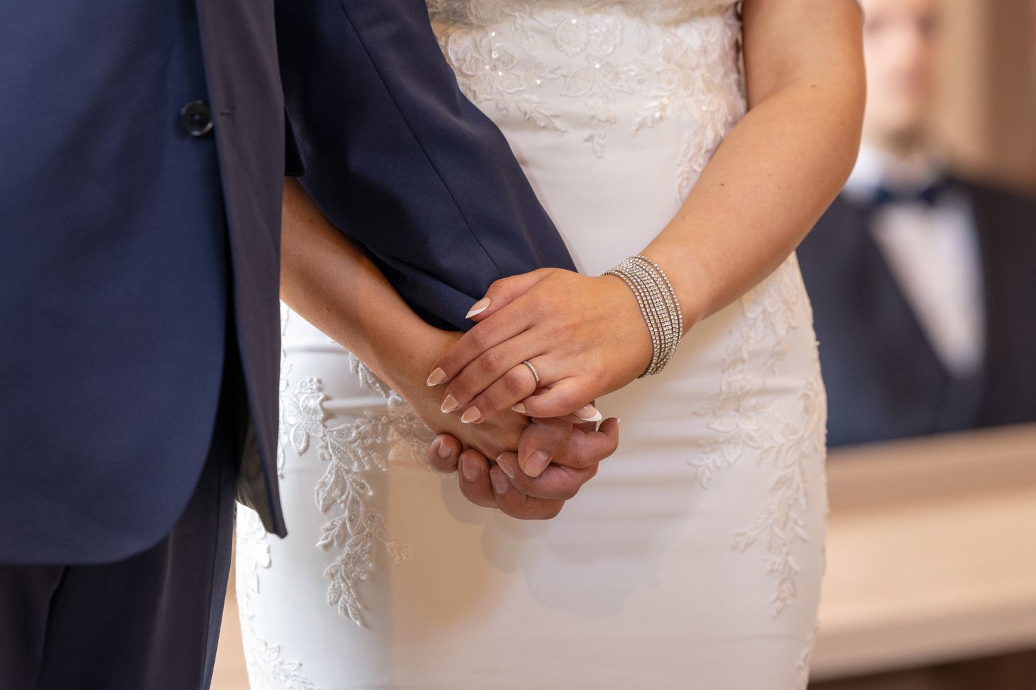 Bride and groom holding hands at wedding ceremony, bride wearing lace dress and diamond bracelet, groom in navy suit.