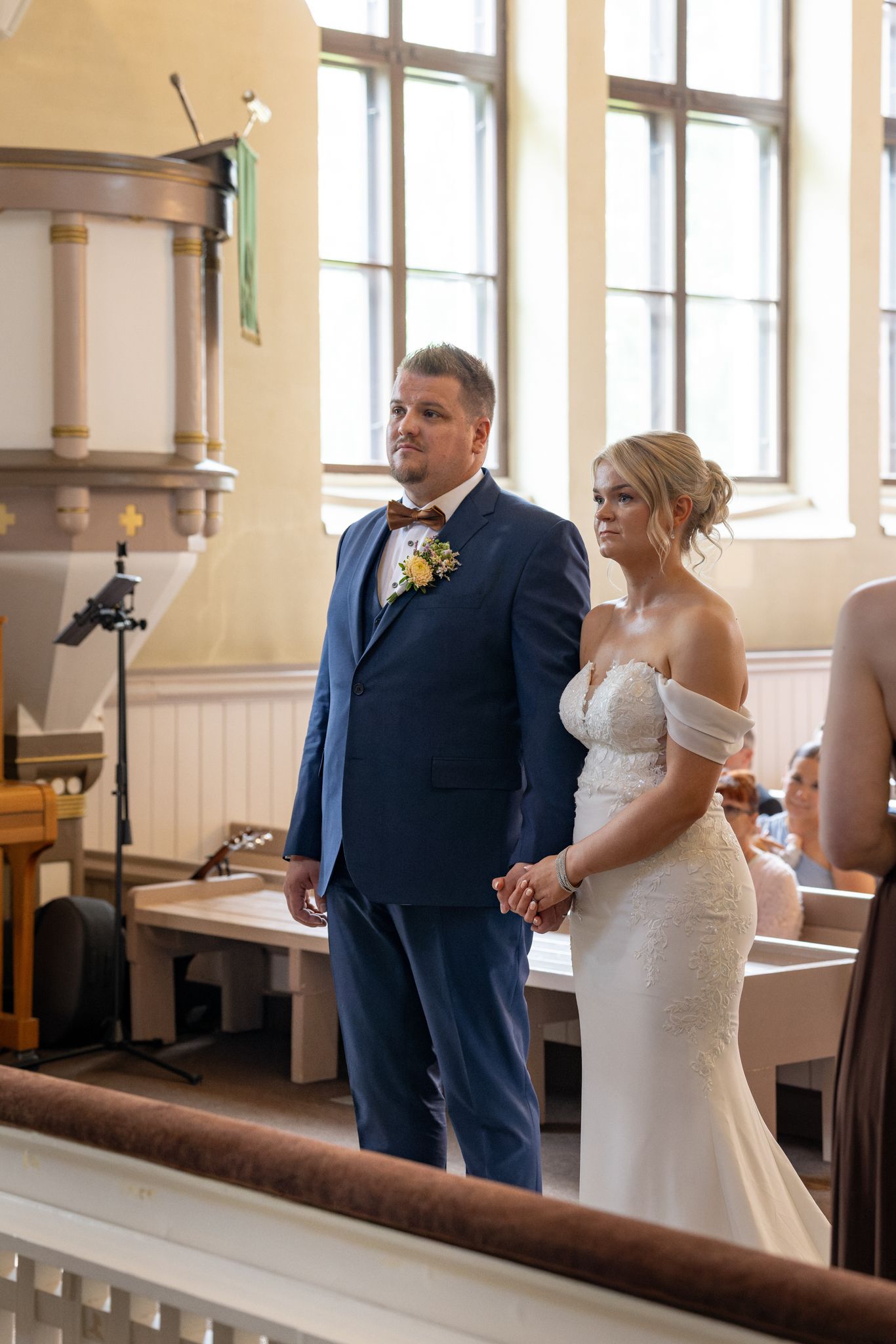 Bride and groom holding hands at church altar, groom in blue suit with boutonniere, bride in off-shoulder white gown.