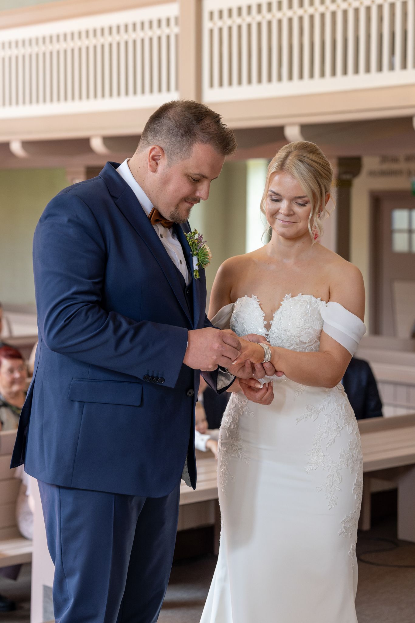 Groom in navy suit places wedding ring on brides finger during ceremony, bride smiles in white off-shoulder gown.