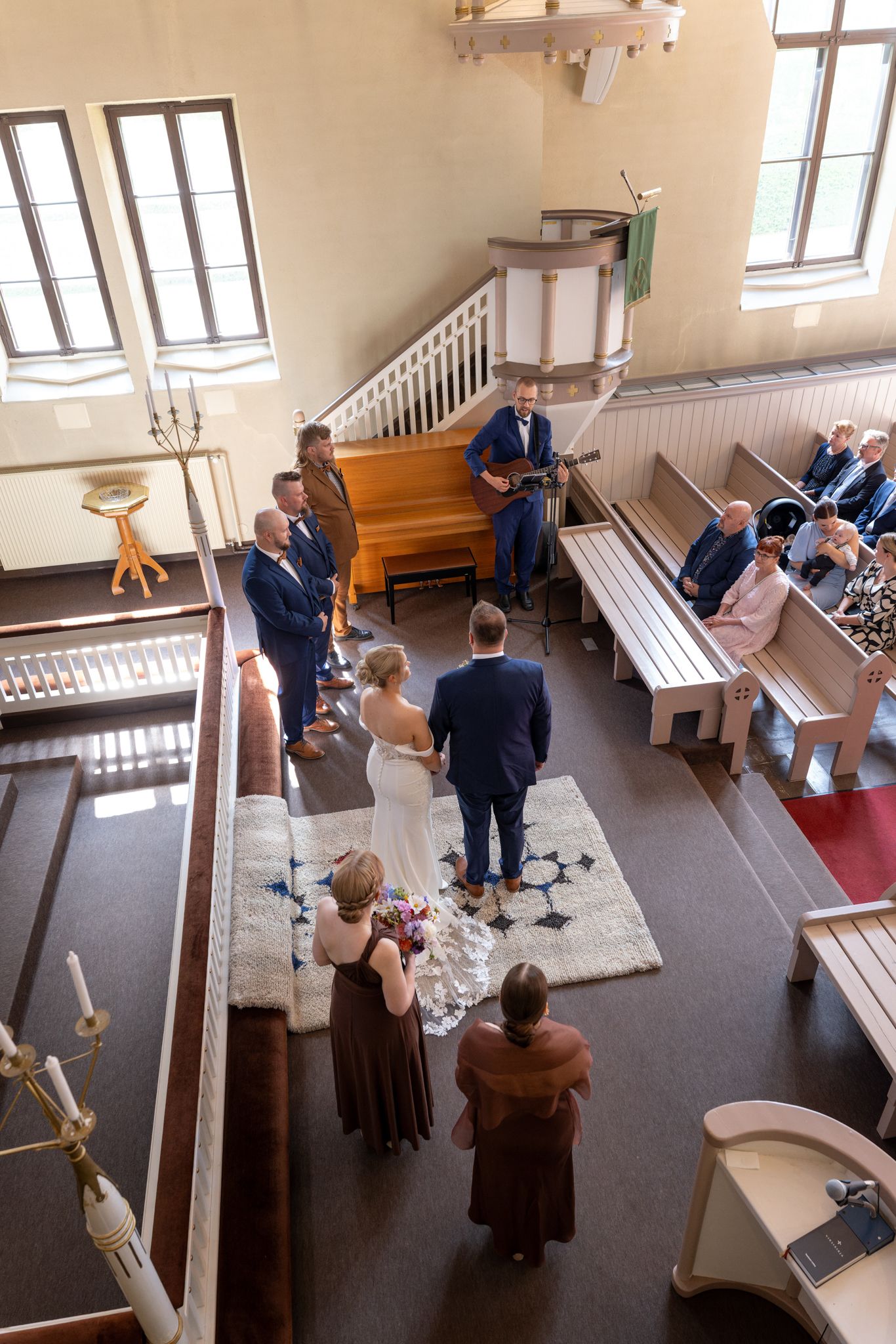 Aerial view of a wedding ceremony inside a small church with bride, groom, and guests seated in wooden pews.