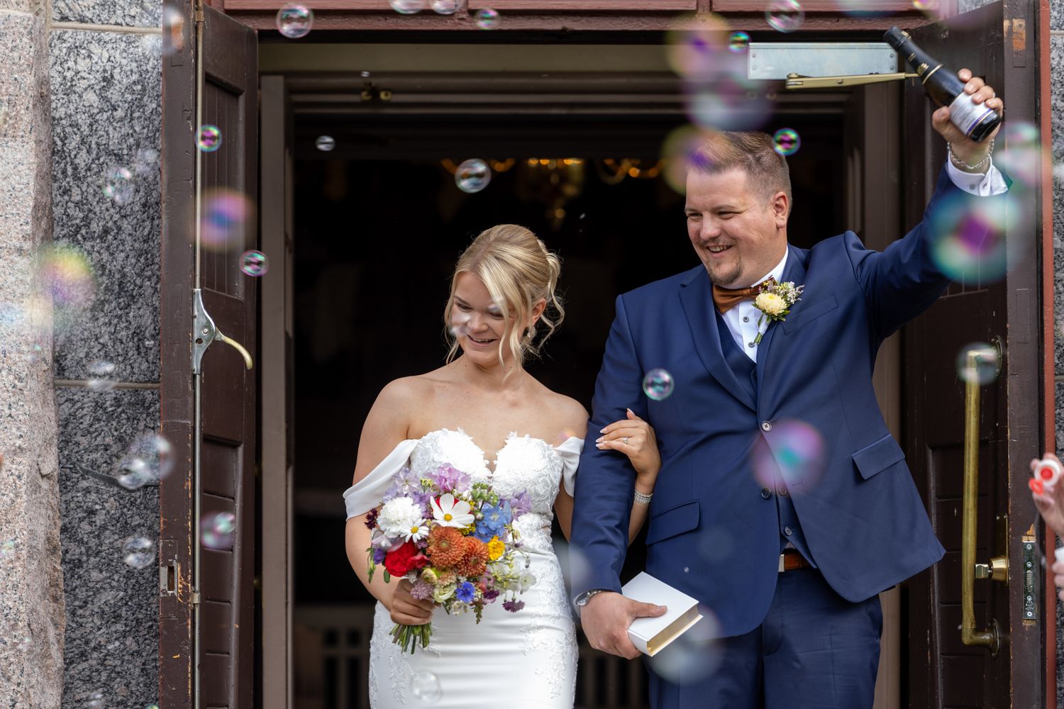Newlyweds exit church doors surrounded by bubbles, bride holding colorful bouquet, groom in navy suit spraying champagne.