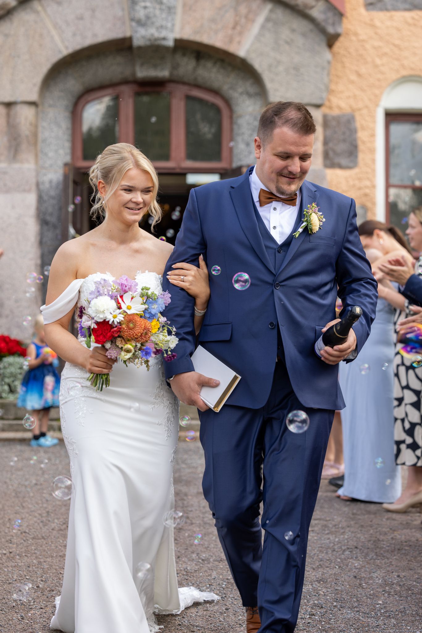 Bride in white gown holding colorful bouquet walks with groom in navy suit amid bubbles after wedding ceremony.