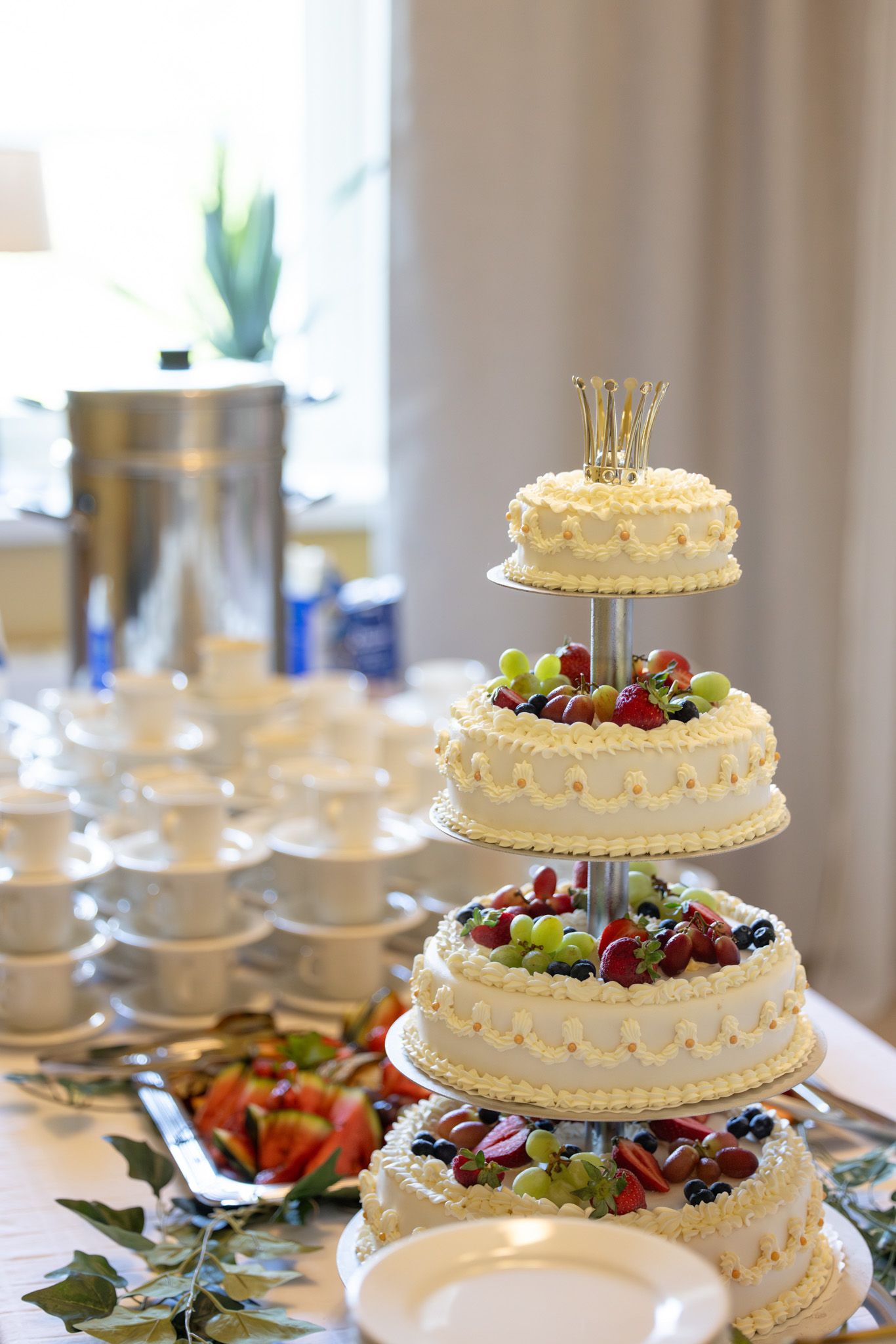Elegant three-tier white wedding cake decorated with fresh fruits and a gold crown topper on a festive dessert table.