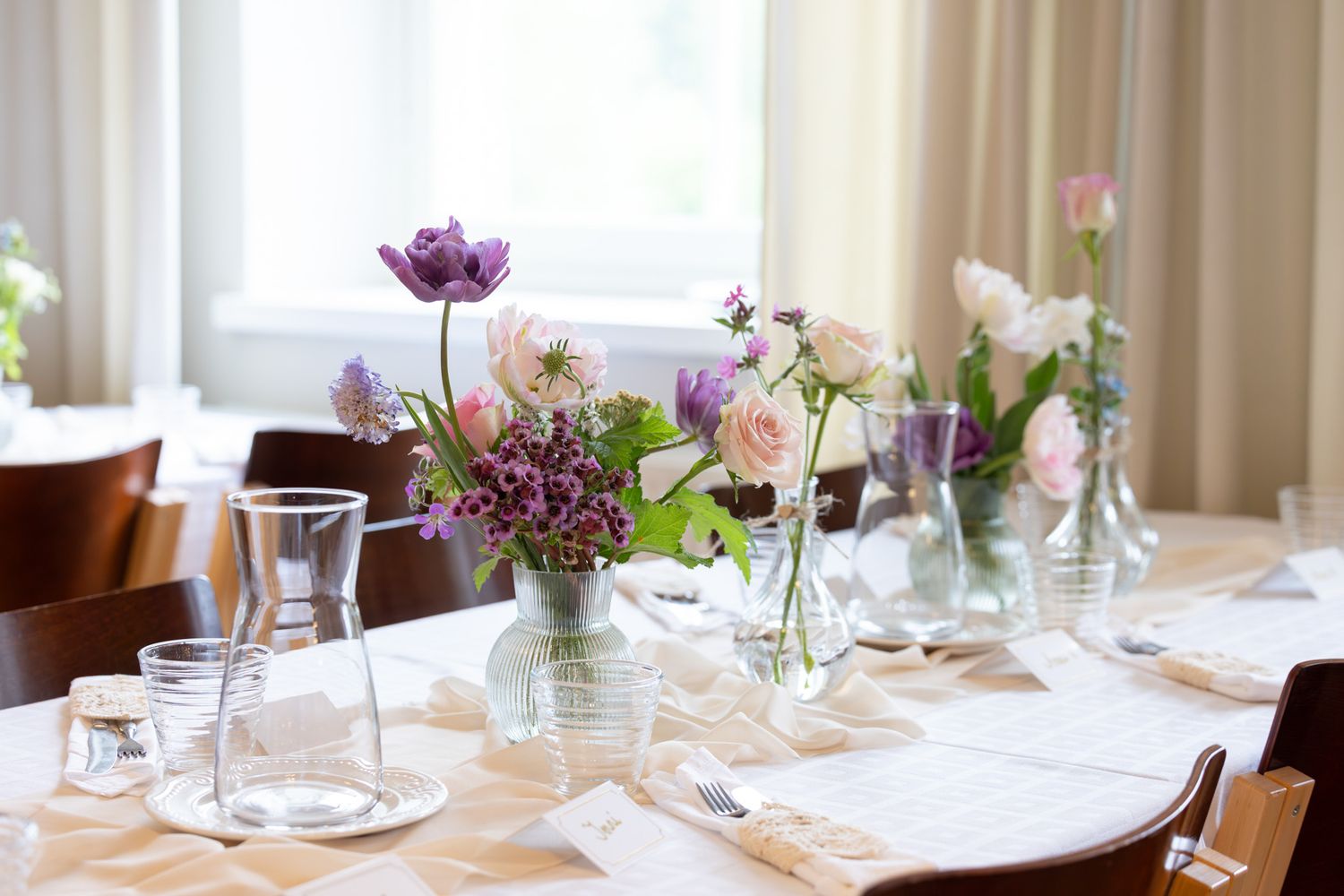 Elegant dining table set with floral centerpieces in glass vases featuring pink roses and purple anemones for a wedding reception.