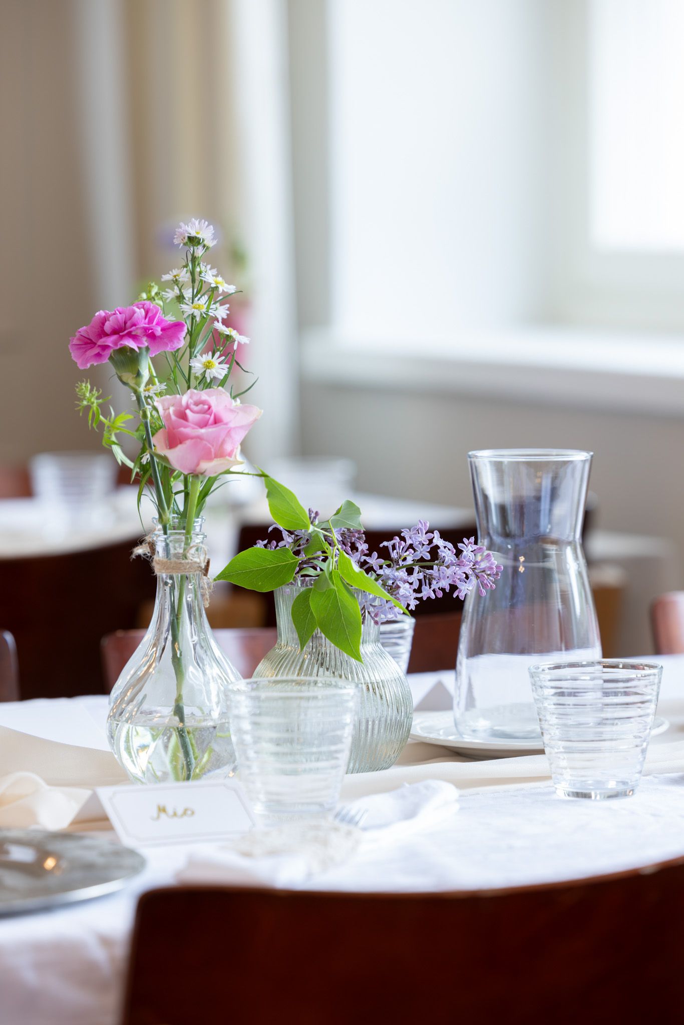 Elegant restaurant table setting with pink roses, wildflowers, and glass carafes on a white tablecloth.