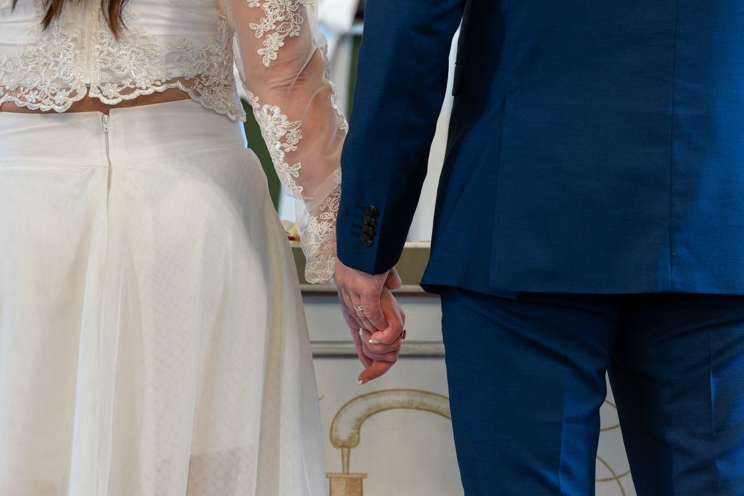 Close-up of couple holding hands, with bride in lace dress and groom in navy suit.