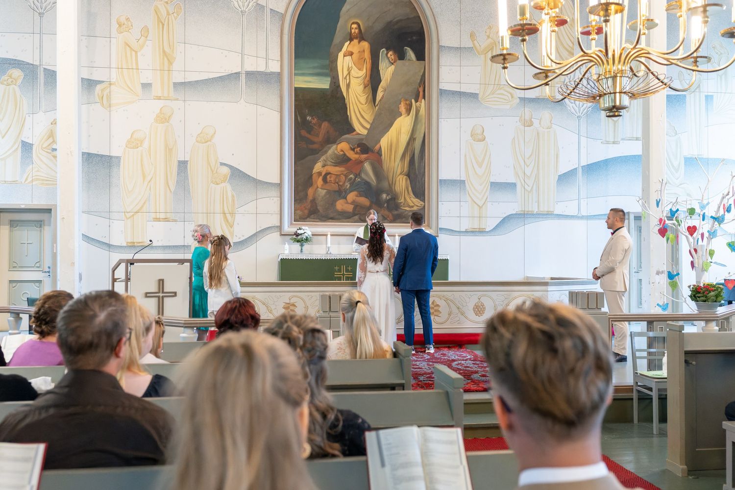 Interior view of a bright white church with chandelier, pews, and religious artwork adorning the walls.