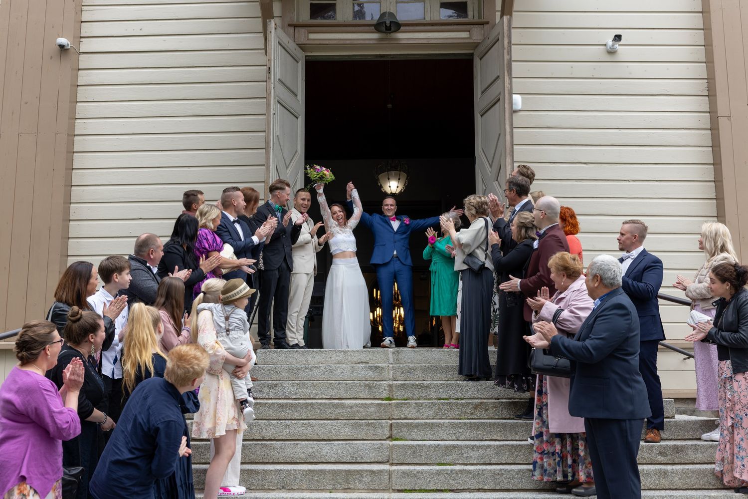 Wedding guests gather outside a beige building entrance during a celebration event.