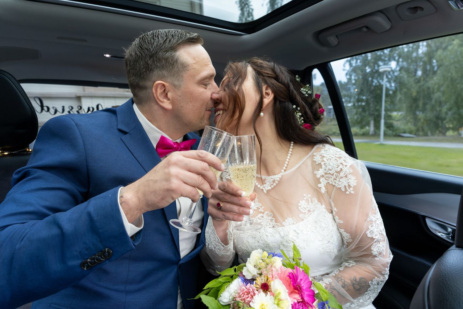 Newlyweds sharing champagne toast in back of car with bridal bouquet of pink flowers.