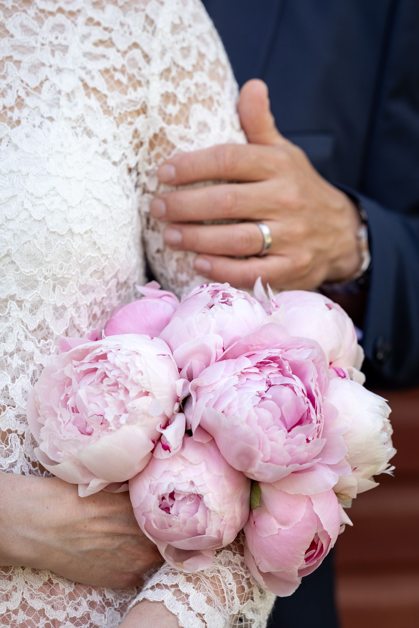 Bride holding pink peony bouquet with groom's hand showing wedding ring on her white lace dress.