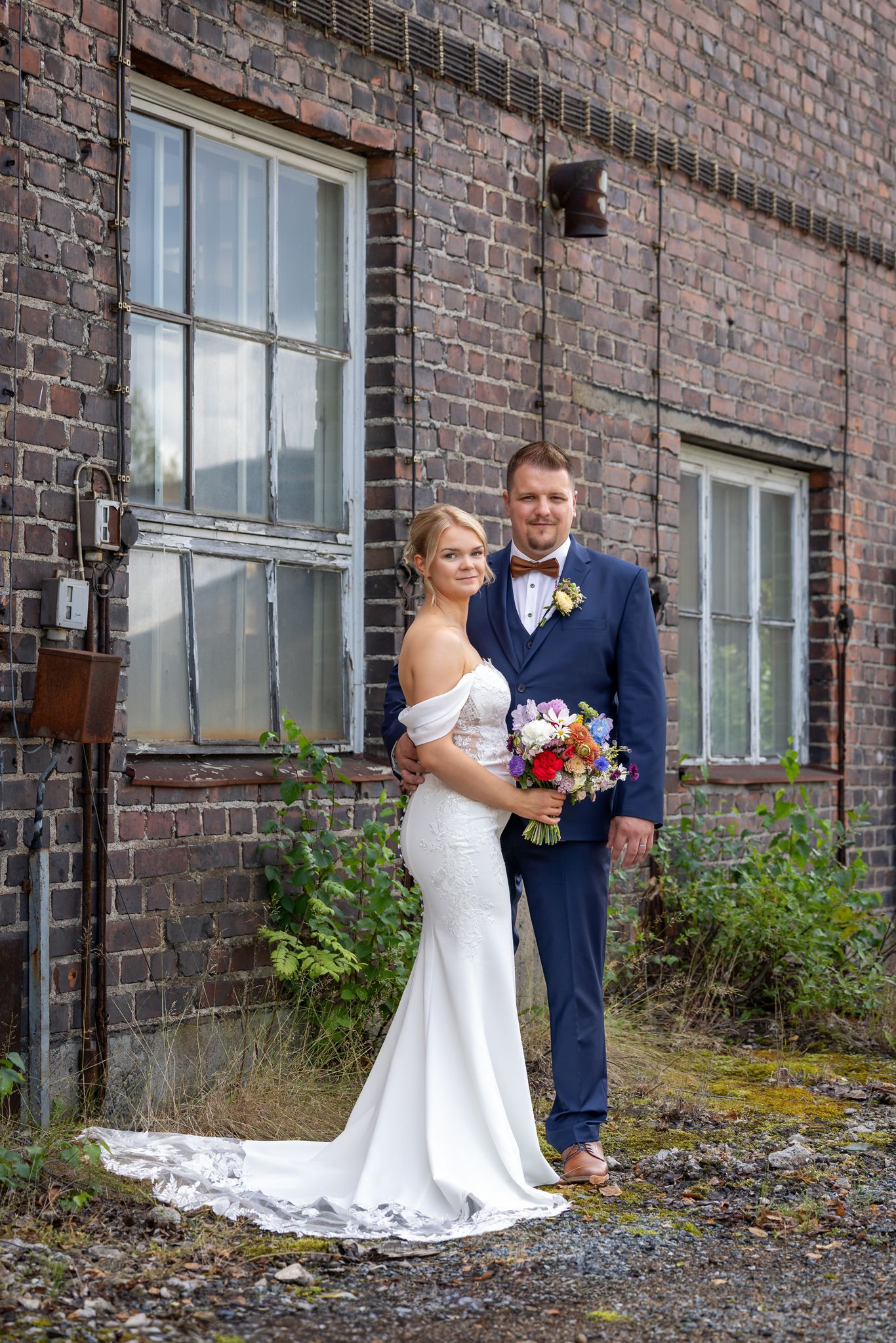 Bride in white off-shoulder gown and groom in navy suit pose with colorful bouquet before rustic brick building.