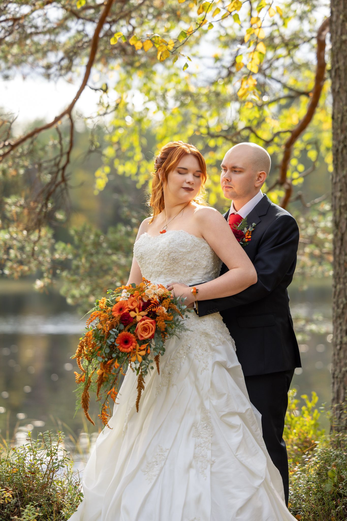 Bride in white gown holding autumn bouquet embraced by groom in black suit near lake in fall forest.