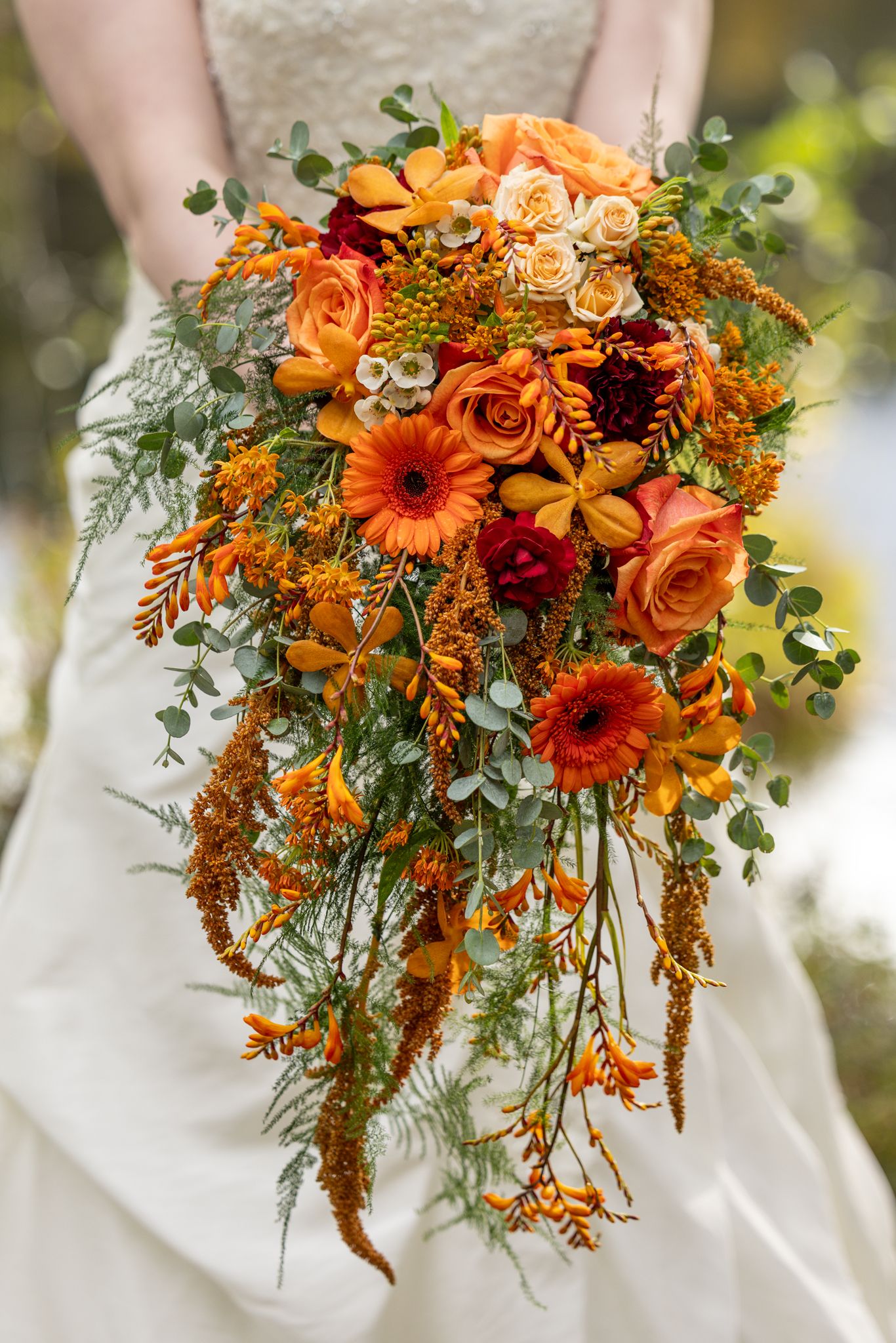 Bride holding a cascading autumn wedding bouquet with orange gerberas, roses, and eucalyptus greenery.