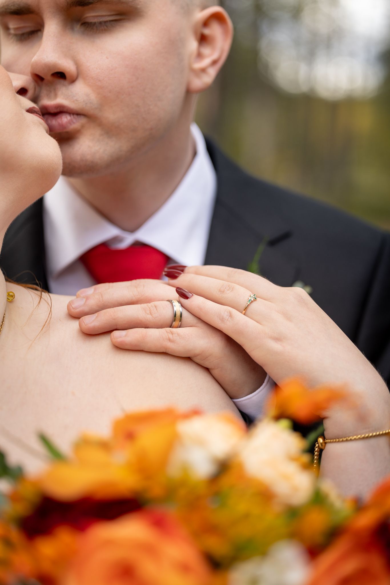 Bride and groom sharing a kiss, displaying wedding rings, with a vibrant autumn bouquet of orange and red flowers in the foreground.
