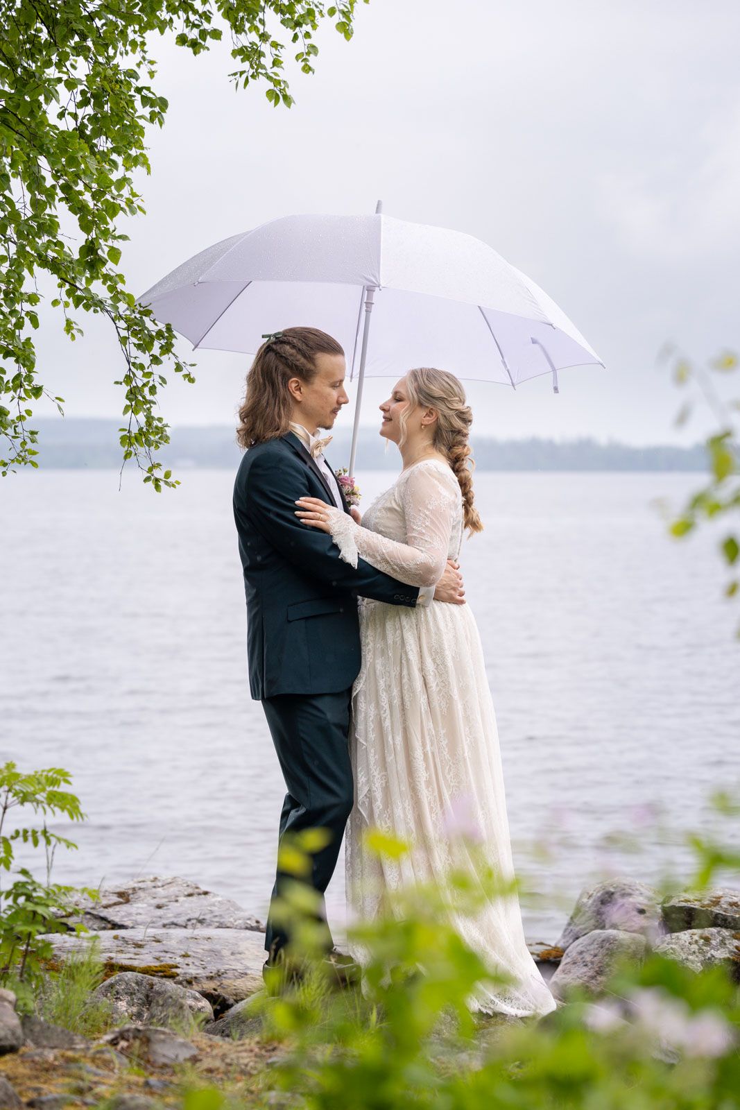 Bride and groom embrace under white umbrella by lakeside on rainy wedding day, wearing elegant white dress and dark suit.