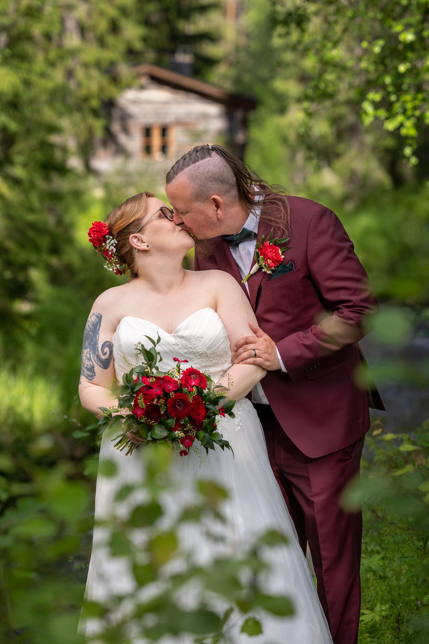 Couple in wedding attire sharing a romantic kiss in a lush garden setting with red floral bouquet.