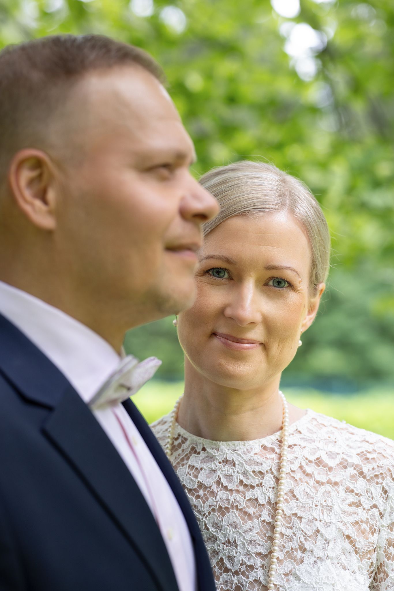 A bride in white lace gazes at her groom in a navy suit outdoors with green trees in the background.