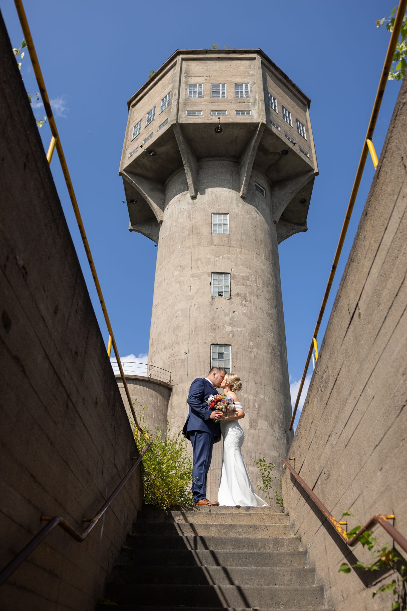 Bride and groom kissing on steps before a historic concrete water tower under a clear blue sky.