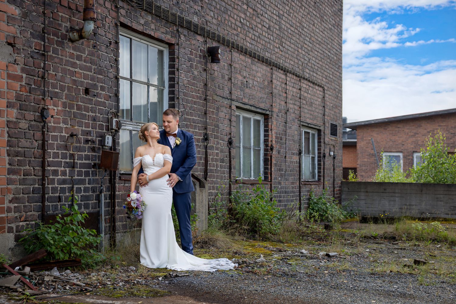 Bride and groom pose romantically against a rustic brick building outdoors on their wedding day.
