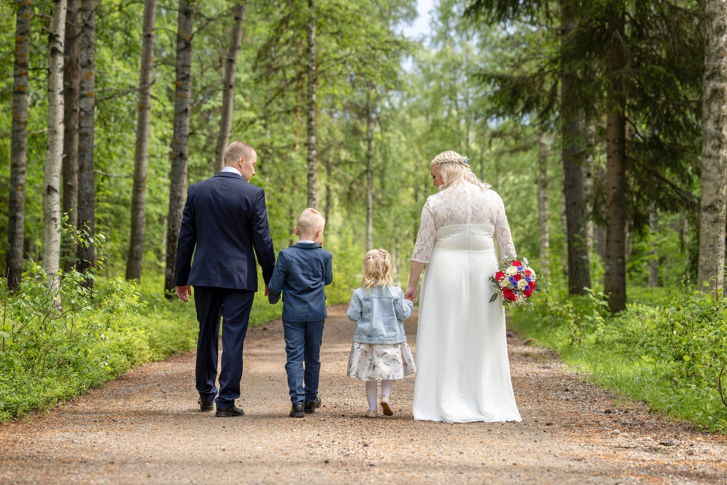 Bride and groom walking with two children on a forest path, bride holding bouquet, wedding day family photo.