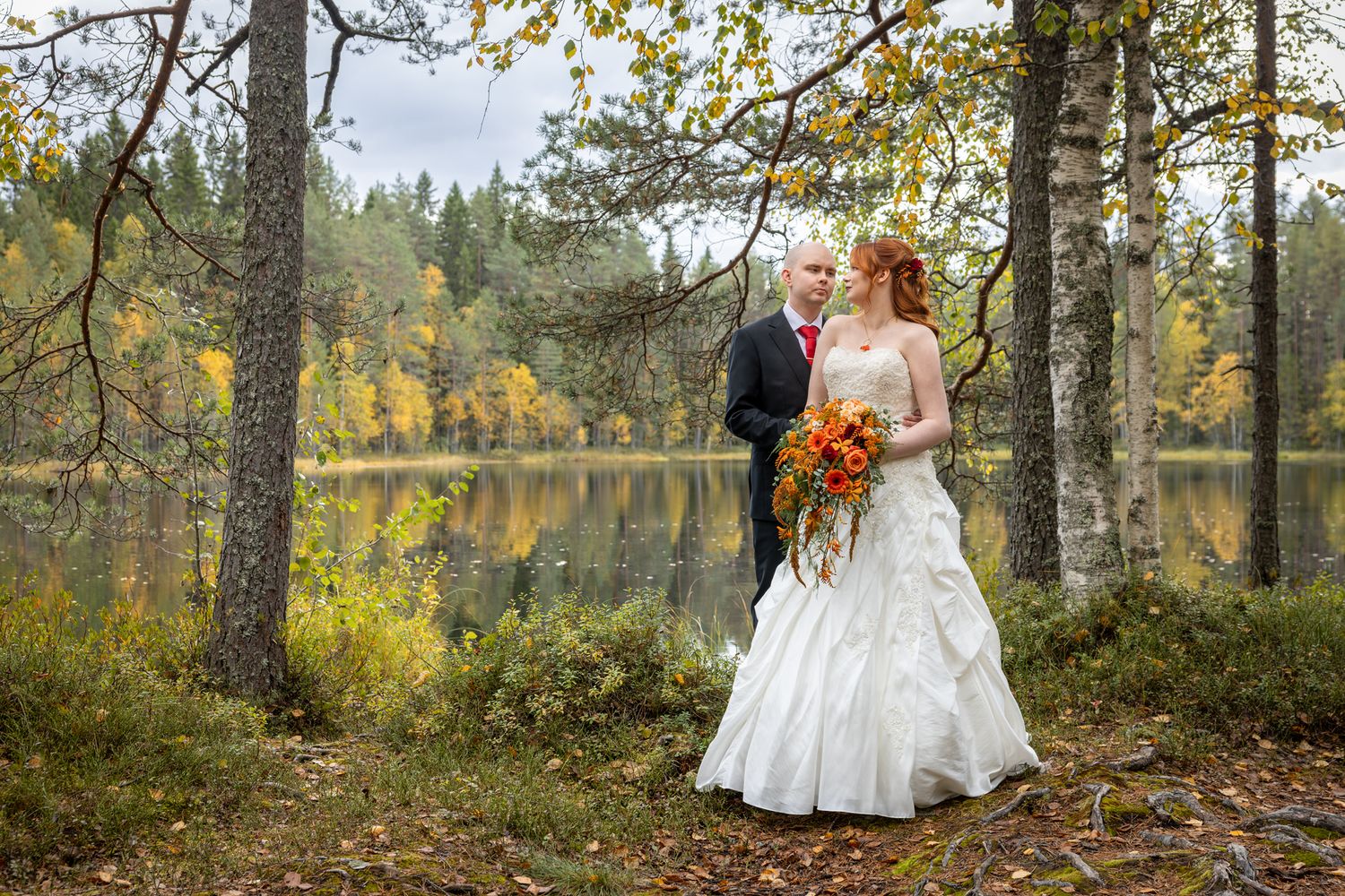 Bride and groom stand among autumn trees beside a serene lake, bride holding a vibrant fall bouquet in a white gown.