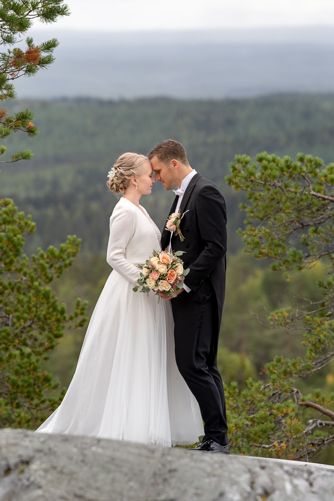 hääpari seisoo kalliolla komeassa vaaramaisemassa. Taustalla taivasta ja metsää.