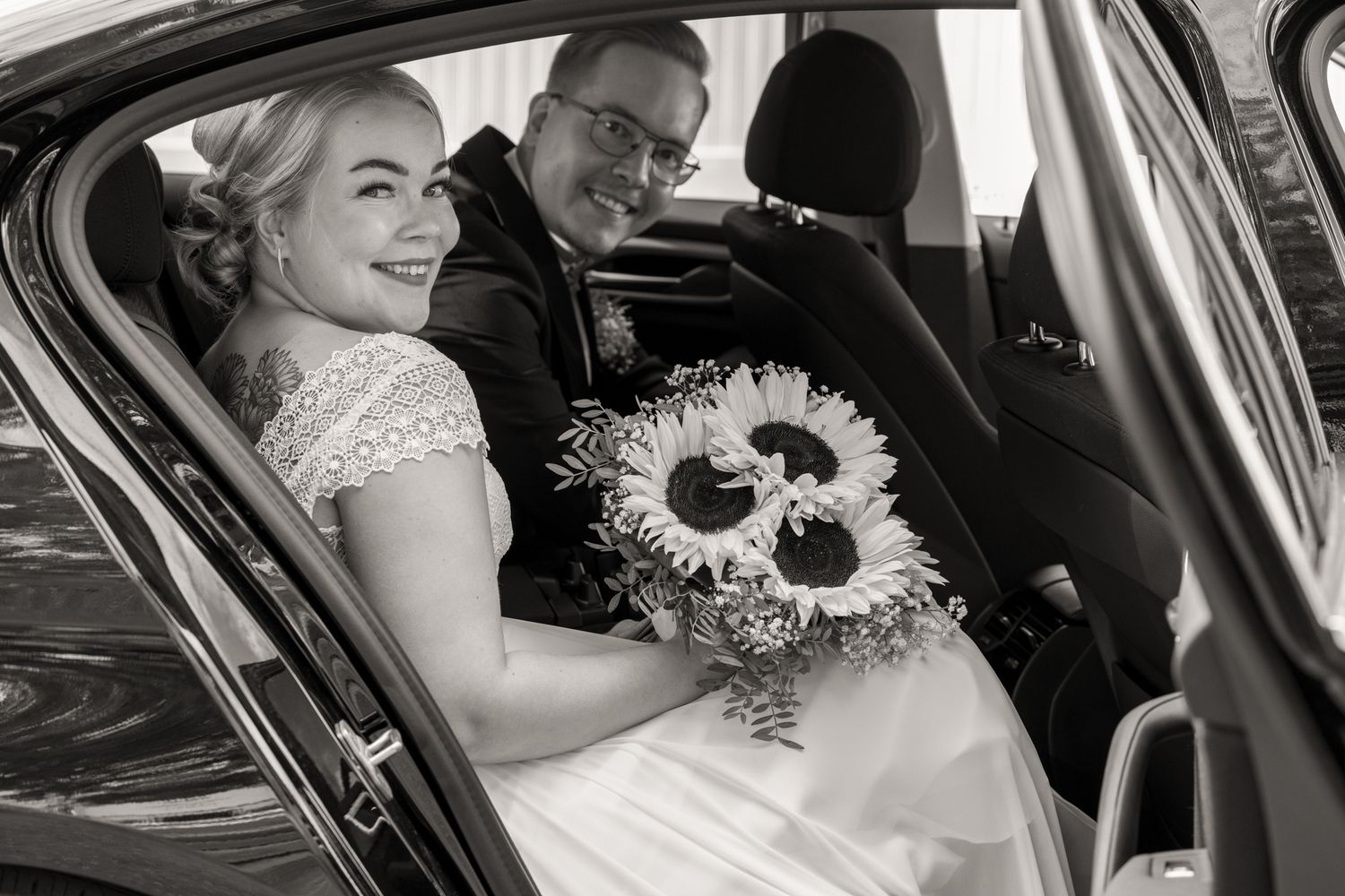 Mustavalkoinen kuva hääautossa istuvasta hääparista valmiina lähtemään kohti hääjuhlaa.