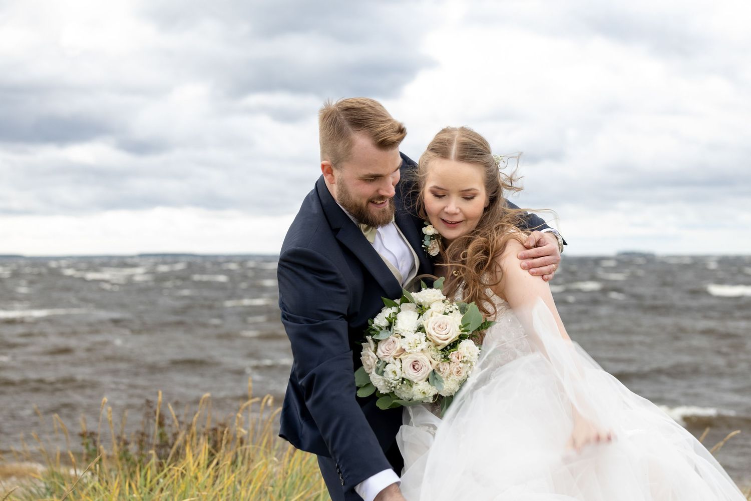Hääpari koettaa taltuttaa tuulessa leijuvaa mekonhelmaa. Taustalla myrskyävä meri.