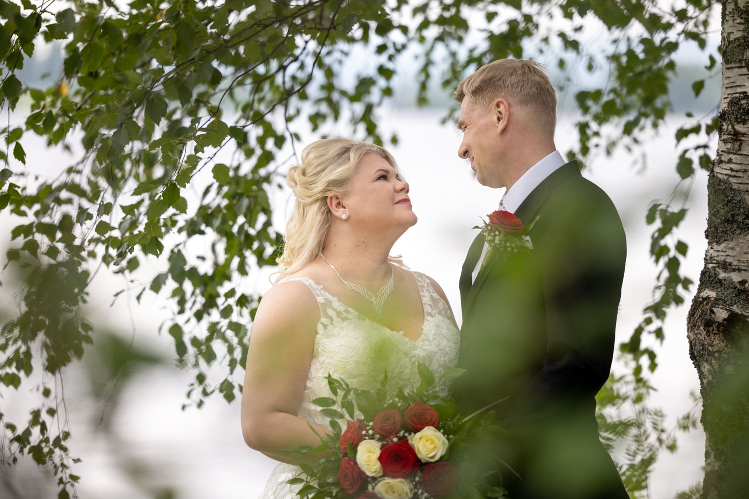 Hääpari seisoo koivun oksien katveessa toisiaan silmiin katsellen. Taustalla järvi.